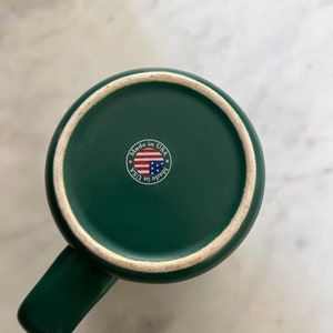 Puede incluir: Una taza de cer&aacute;mica verde con asa. La parte inferior de la taza tiene una pegatina que dice "Made in USA" con una peque&ntilde;a bandera estadounidense.