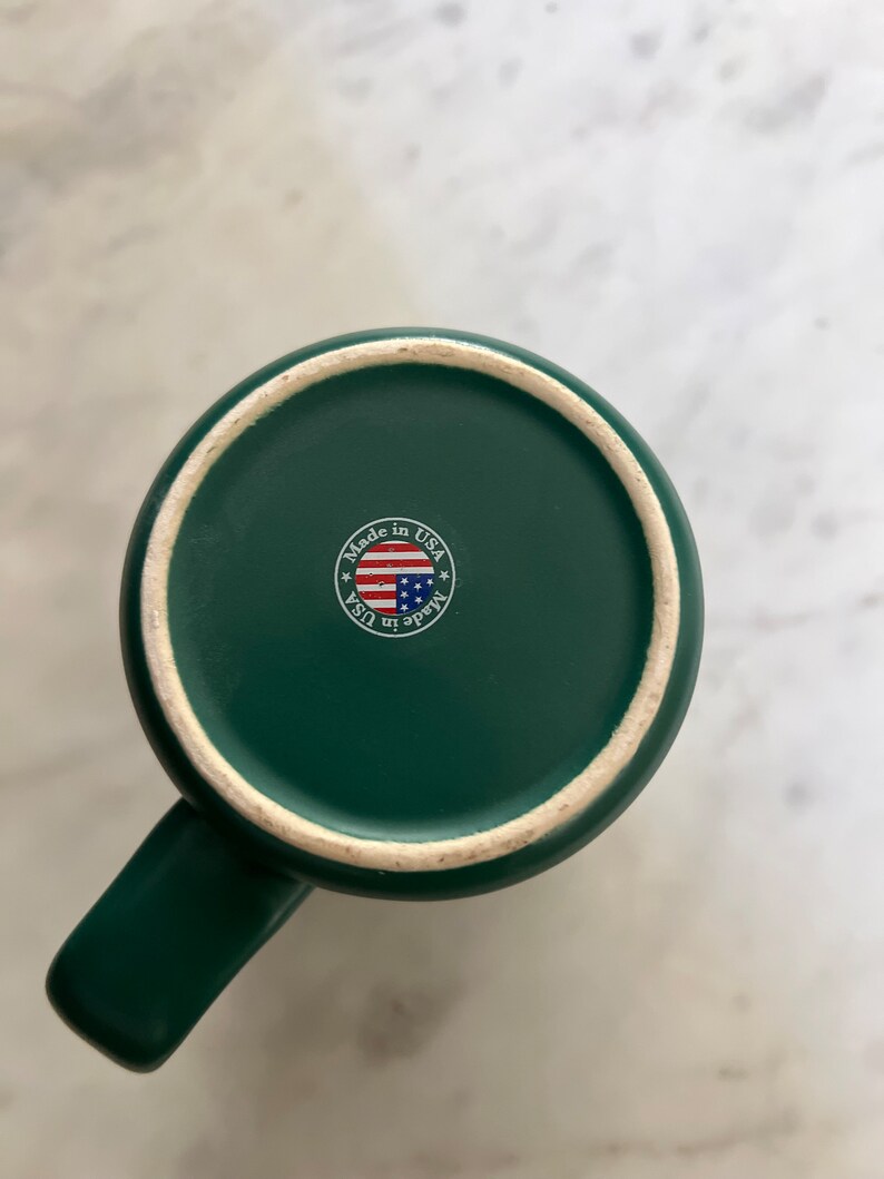 Puede incluir: Una taza de cer&aacute;mica verde con asa. La parte inferior de la taza tiene una pegatina que dice "Made in USA" con una peque&ntilde;a bandera estadounidense.
