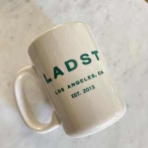 Puede incluir: Taza de cer&aacute;mica blanca con un logotipo verde que dice "LADST LOS ANGELES, CA EST. 2015"