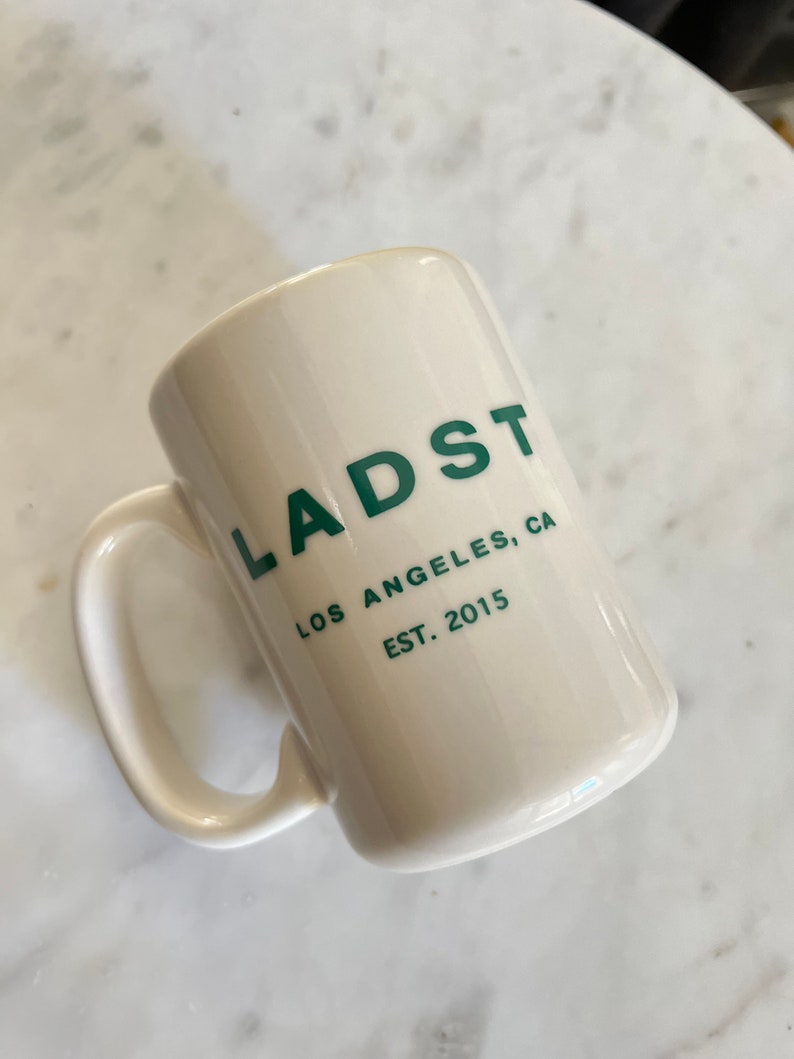 Puede incluir: Taza de cer&aacute;mica blanca con un logotipo verde que dice "LADST LOS ANGELES, CA EST. 2015"