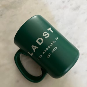 Puede incluir: Una taza de cer&aacute;mica verde con el texto "LADST LOS ANGELES, CA EST. 2015" impreso en blanco.