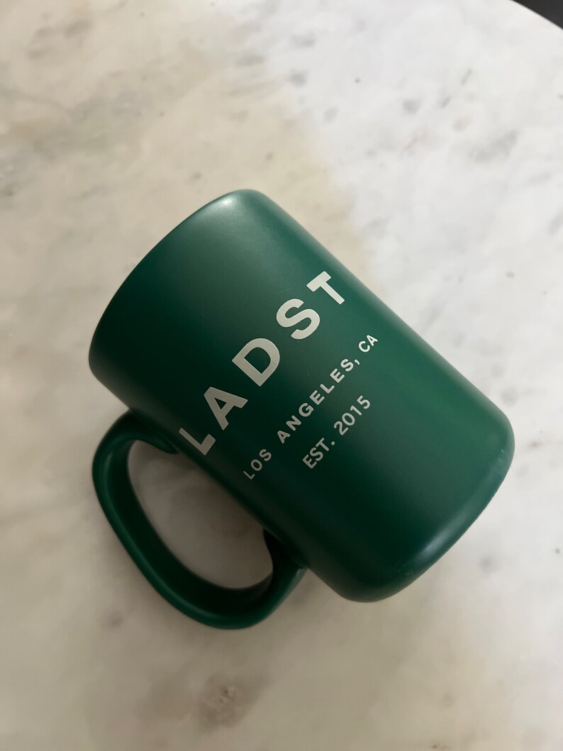 Puede incluir: Una taza de cer&aacute;mica verde con el texto "LADST LOS ANGELES, CA EST. 2015" impreso en blanco.