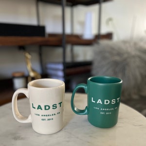 Puede incluir: Dos tazas de cer&aacute;mica con el texto "LADST LOS ANGELES, CA EST. 2015" impreso en ellas. Una taza es blanca con texto verde y la otra es verde con texto blanco.