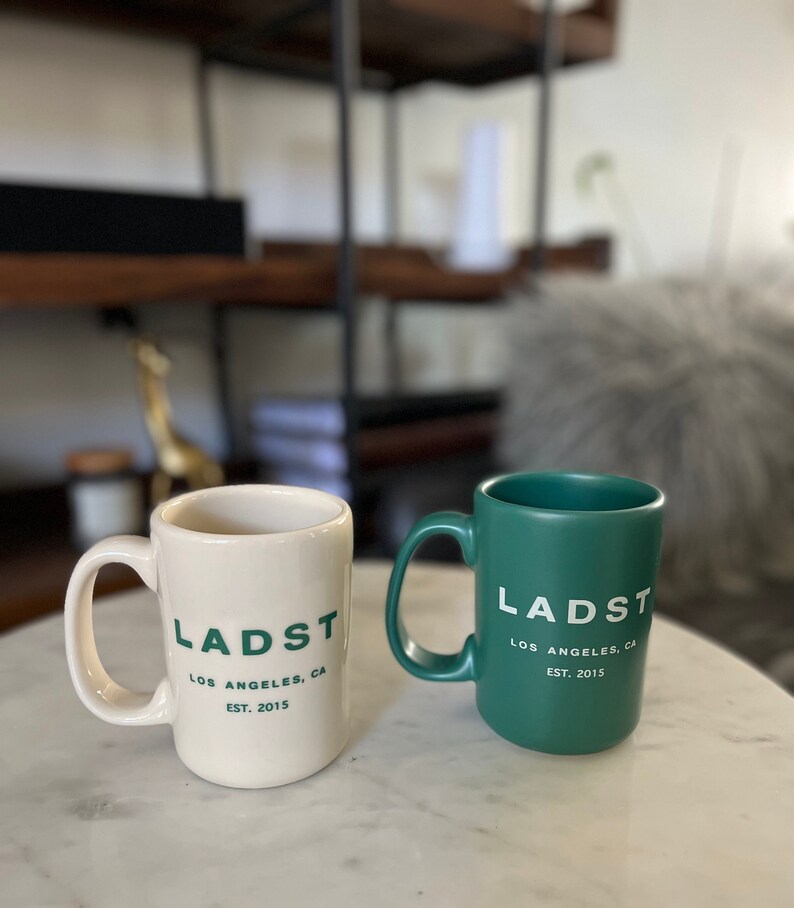 Puede incluir: Dos tazas de cer&aacute;mica con el texto "LADST LOS ANGELES, CA EST. 2015" impreso en ellas. Una taza es blanca con texto verde y la otra es verde con texto blanco.