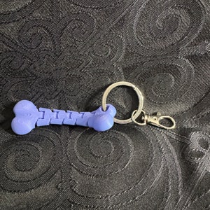 Puede incluir: Un llavero de plástico morado con forma de hueso con un anillo plateado y un cierre plateado.