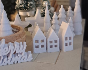 May include: White miniature houses with windows, a small white Christmas tree, and a sign that says "Merry Christmas" on a white surface.