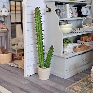 Puede incluir: Un cactus en miniatura en una maceta color crema. El cactus tiene un tallo alto, verde y espinoso y otro más pequeño similar. La maceta está sobre un suelo de madera gris. Un mueble de cocina en miniatura está en el fondo.