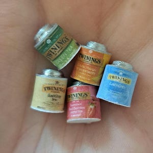 May include: Five small, silver, cylindrical tea tins with colorful labels. The labels feature the brand name "Twinings" and different tea flavors, including Green Tea, Earl Grey, Lemon & Mango,  Strawberry & Raspberry, and Lady Grey.