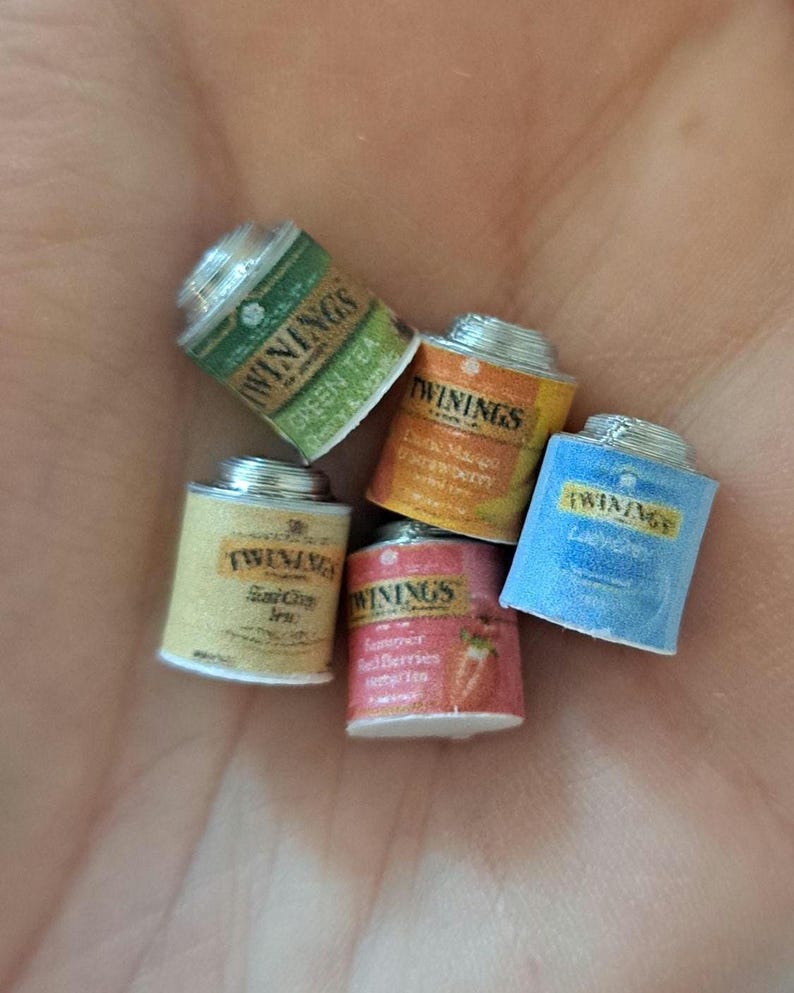 May include: Five small, silver, cylindrical tea tins with colorful labels. The labels feature the brand name "Twinings" and different tea flavors, including Green Tea, Earl Grey, Lemon & Mango,  Strawberry & Raspberry, and Lady Grey.