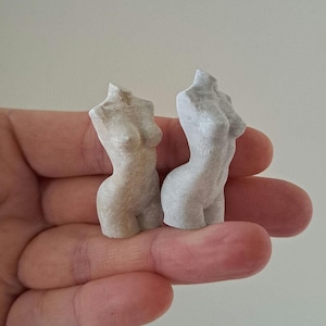 May include: Two small, off-white, sculpted torso figurines. The figures are smooth with defined curves and details. The figurines are held in a hand, showcasing their size and texture. The sculptures are a light, neutral color.