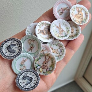 May include: A collection of miniature decorative plates featuring Easter bunny designs. The plates have various pastel color schemes, including pink, green, and white, with floral and striped borders. The bunny illustrations vary in style and detail.