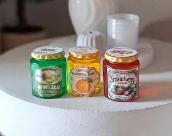 Può includere: Tre barattoli di vetro in miniatura riempiti di marmellata traslucida verde, gialla e rossa. Ogni barattolo presenta un coperchio color oro e un'etichetta di carta. Le etichette mostrano illustrazioni di kiwi, arancia e fragola con il testo Kiwi, Orange e Strawberry.