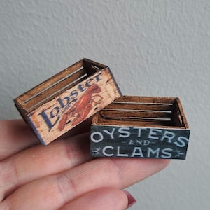 Może przedstawiać: Dwie miniaturowe drewniane skrzynki z otwartą górą. Jedna skrzynka ma ciemnobrązowe wykończenie z napisem Lobster wydrukowanym na niebiesko oraz ilustracją czerwonego homara. Druga skrzynka ma ciemnoszary przedni panel z napisem Oysters and Clams w kolorze białym.