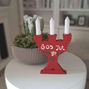 Könnte beinhalten: Ein roter hölzerner Kerzenhalter mit drei weißen Stabkerzen. Die Vorderseite des Halters ist handbemalt mit dem weißen Schriftzug God Jul und einer kleinen weißen Blume mit grünen Blättern. Das Objekt hat einen breiten Fuß und drei Kerzenhalter.