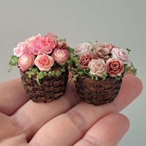 May include: Two miniature woven baskets filled with pink and red roses and carnations. The flowers are surrounded by green foliage. The baskets are held in a hand.