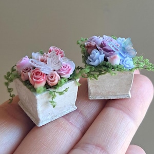 May include: Two miniature flower boxes, each filled with detailed, colorful roses and greenery. One box features pink and white roses with a butterfly accent, while the other has blue and purple roses with a butterfly. The boxes are held in a hand.