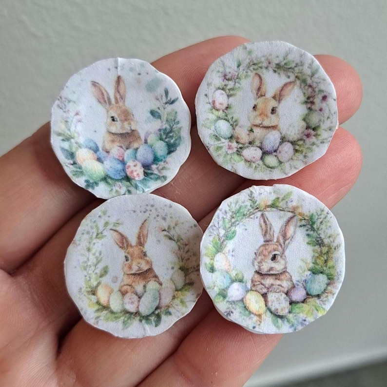 May include: Four small, round Easter-themed decorations. Each features a watercolor illustration of a brown bunny surrounded by colorful Easter eggs and greenery. The decorations are on a white background.