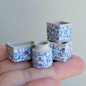 Könnte beinhalten: Miniatur-Keramikvasen und -Pflanzgefäße mit blauem Blumenmuster. Das Set besteht aus einer kleinen Vase und drei quadratischen Pflanzgefäßen. Die Ränder der Pflanzgefäße sind hellgrau eingefasst. Geeignet für ein Puppenhaus oder eine Miniaturgarten.