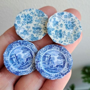 Op de afbeelding: Vier miniatuur keramische borden. Twee borden hebben een blauw bloemmotief op een crèmekleurige achtergrond. De andere twee borden hebben een blauw-wit landschapsontwerp met een decoratieve rand. Ideaal voor poppenhuizen of knutselprojecten.
