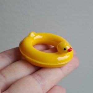 以下が含まれることがあります： 赤いクチバシと黒い目を持つ、ミニチュアの黄色のラバーダック浮き輪。小さくて丸い浮遊装置は明るい黄色で、側面にアヒルの頭が付いています。