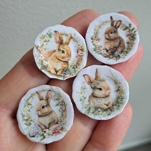 May include: Four small, round decorative images featuring watercolor illustrations of bunnies surrounded by floral wreaths. The bunnies have varying shades of brown fur and are set against a white background. The floral wreaths include pink, green, and yellow flowers.