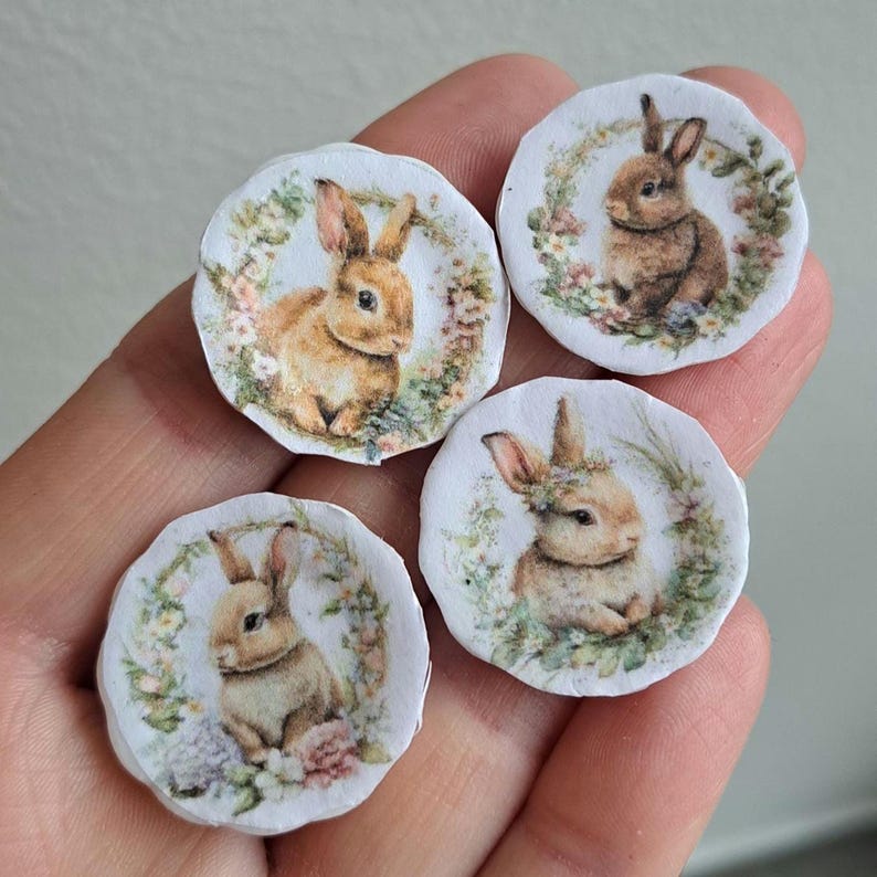 May include: Four small, round decorative images featuring watercolor illustrations of bunnies surrounded by floral wreaths. The bunnies have varying shades of brown fur and are set against a white background. The floral wreaths include pink, green, and yellow flowers.