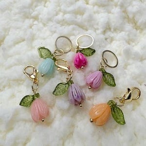 Peut inclure: Plusieurs breloques en forme de fleurs colorées. Chaque breloque est dotée d'un fermoir et d'un anneau dorés, avec une fleur translucide dans les tons de rose, violet, bleu et orange, et des feuilles vertes.