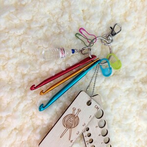 Peut inclure: Un jauge à crochet en bois avec une règle d'un côté et une rangée de trous pour différentes tailles d'aiguilles de crochet de l'autre. La jauge est attachée à un porte-clés avec un petit charme de bouteille d'eau, quelques épingles de sûreté colorées et trois aiguilles à crochet en rouge, orange et bleu.