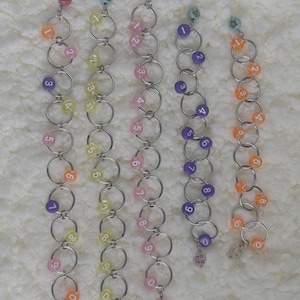 May include: A selection of handmade beaded keychains. Each keychain has silver rings with colourful beads and numbered accents. The beads are in shades of pink, orange, yellow, and purple. Some keychains have smiley face charms.