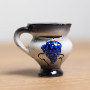 May include: A small white ceramic pitcher with a black rim and handle. The pitcher has a blue and brown grape design on the side.