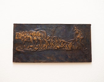 May include: A bronze plaque depicting a scene of a farmer plowing a field with a team of oxen. The plaque is rectangular and has a dark brown patina. The oxen are pulling a plow, and the farmer is walking behind them. The scene is detailed and realistic.