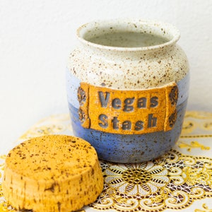 Puede incluir: Un tarro de cerámica con un esmalte blanco moteado y una franja azul y amarilla. El tarro tiene una tapa de corcho y dice "Vegas Stash" en la parte delantera.