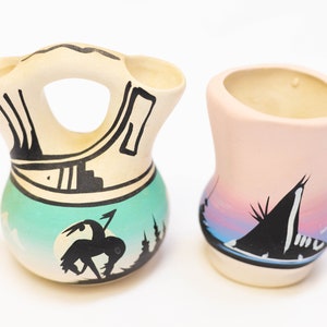 May include: Two small ceramic pottery vases with hand-painted designs. One vase is a traditional Southwestern style with a turquoise and white background and black geometric patterns. The other vase is a light pink with a black silhouette of a teepee and a sunset scene.