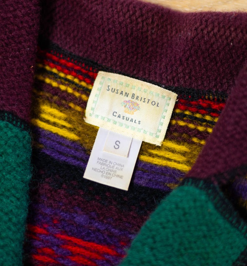 May include: A close-up of a brown, green, purple, yellow, and red striped fleece jacket with a tag that reads "Susan Bristol Casuals" and "S".