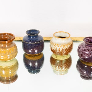 Peut inclure: Quatre petits vases en céramique faits à la main. Chaque vase présente une palette de couleurs unique : marron et jaune, bleu foncé, crème et marron, et violet tacheté. Les vases ont une forme arrondie avec une petite ouverture.