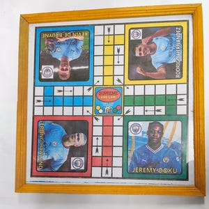May include: A framed Ludo board game featuring four players: Kevin De Bruyne, Rodrigo Hernandez, Erling Haaland, and Jeremy Doku. The board has a colorful design with the text "SUPREME LUDO GAME" in the center. The frame is a light brown color.