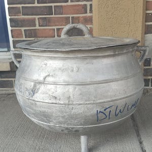 Puede incluir: Una olla grande y redonda de aluminio con tapa y dos asas laterales. La olla tiene una sola pata y está sobre una superficie de hormigón. La olla tiene algunas escrituras en el lateral.