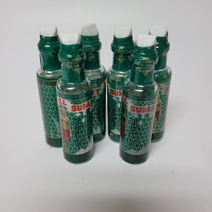 Puede incluir: Un conjunto de siete botellas de vidrio con tapas y etiquetas verdes. Las etiquetas presentan una figura con pantalones cortos rojos y el texto "NIGERIA SUDAN". Las botellas están llenas de un líquido transparente.