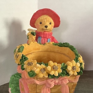 May include: A ceramic planter shaped like a wagon with a teddy bear wearing a red hat and a pink sweater. The wagon is decorated with yellow flowers and a pink ribbon.