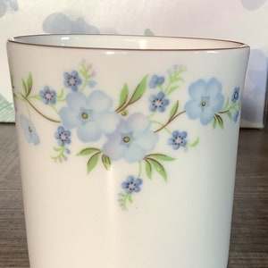 May include: White ceramic tumbler with a floral design. The tumbler features a delicate pattern of light blue flowers and green leaves. The rim is trimmed with a thin brown line. The design is repeated around the tumbler.