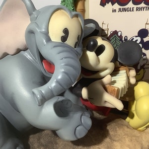Puede incluir: Una figurita de elefante gris con una boca roja y una oreja grande est&aacute; sentada junto a una figurita de Mickey Mouse tocando un acorde&oacute;n. El fondo es un p&oacute;ster con el texto "Mickey Mouse in Jungle Rhythm".