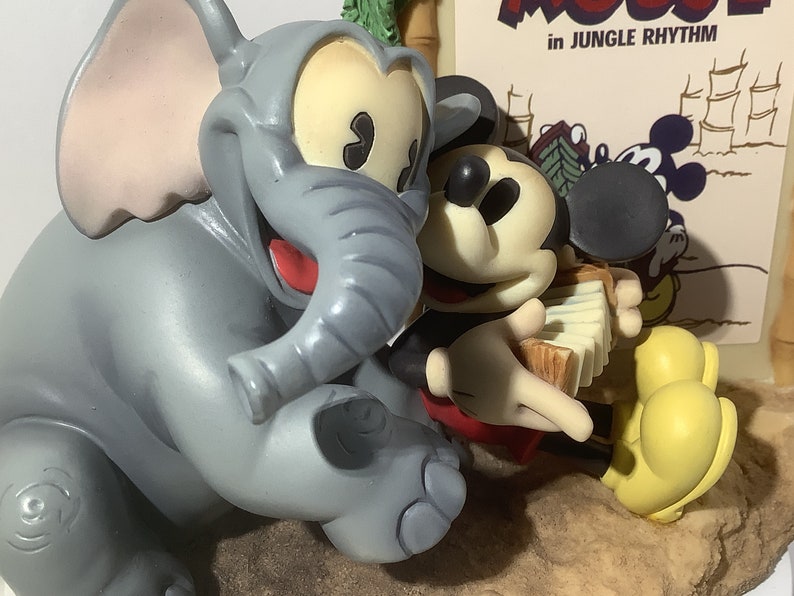 Puede incluir: Una figurita de elefante gris con una boca roja y una oreja grande est&aacute; sentada junto a una figurita de Mickey Mouse tocando un acorde&oacute;n. El fondo es un p&oacute;ster con el texto "Mickey Mouse in Jungle Rhythm".