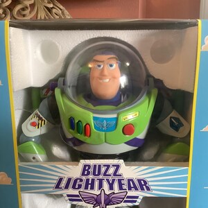 Op de afbeelding: Buzz Lightyear actiefiguur in de originele doos. Het figuur draagt een groen en wit ruimtepak met een doorzichtige helm. De doos is blauw met witte wolken en de tekst "Buzz Lightyear" in een gestileerd lettertype.