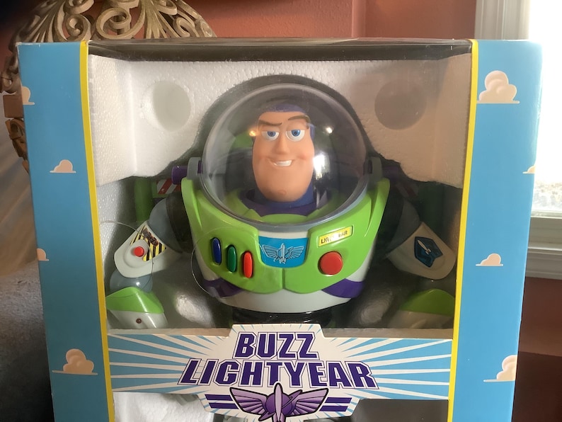 Op de afbeelding: Buzz Lightyear actiefiguur in de originele doos. Het figuur draagt een groen en wit ruimtepak met een doorzichtige helm. De doos is blauw met witte wolken en de tekst "Buzz Lightyear" in een gestileerd lettertype.
