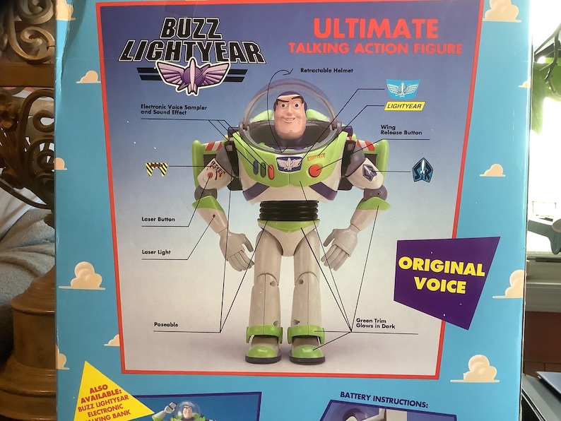 Op de afbeelding: Een doos van de Buzz Lightyear Ultimate Talking Action Figure. De doos toont een gedetailleerde afbeelding van de Buzz Lightyear actiefiguur, met labels die functies benadrukken zoals de intrekbare helm, de laserknop en de vleugelontgrendelingsknop. De doos bevat ook de tekst "Original Voice".