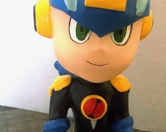 May include: A figurine of a cartoon character with a blue helmet and yellow accents. The character wears a black suit with blue gloves and boots. The figurine has green eyes and a red emblem on the chest.