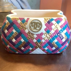 May include: A decorative ceramic handbag-shaped container with a woven pattern in pink, purple, teal, gold, and white. The container has a white interior and a gold emblem on the front.