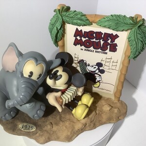 Puede incluir: Una figurita de cer&aacute;mica de Mickey Mouse tocando un acorde&oacute;n con un elefante gris a su lado. La figurita est&aacute; colocada sobre un fondo de marco de madera con un cartel vintage que dice "Mickey Mouse in Jungle Rhythm".