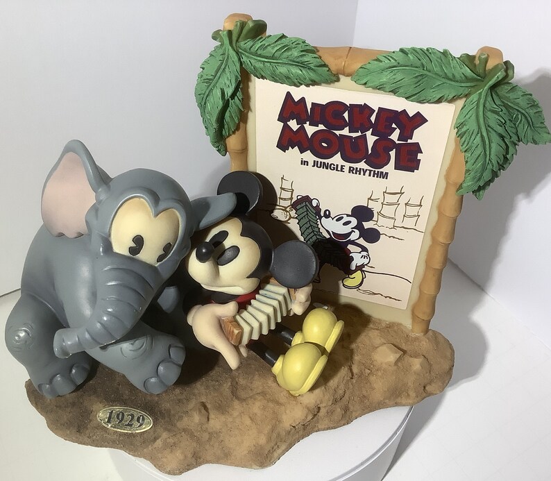 Puede incluir: Una figurita de cer&aacute;mica de Mickey Mouse tocando un acorde&oacute;n con un elefante gris a su lado. La figurita est&aacute; colocada sobre un fondo de marco de madera con un cartel vintage que dice "Mickey Mouse in Jungle Rhythm".