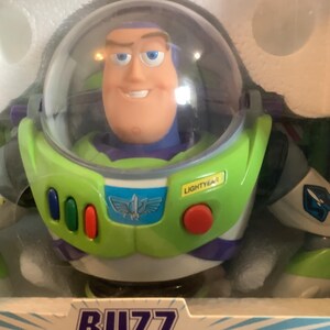 Op de afbeelding: Een Buzz Lightyear actiefiguur in de originele verpakking. De figuur heeft een groen en wit ruimtepak met een heldere helm, een lachend gezicht en het Lightyear-logo. De verpakking is geel en wit.